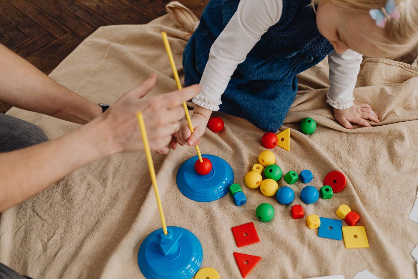A toddler playing with stacking toys on a blanket while an adult helps